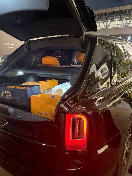 The image shows a black car with its trunk open, revealing several boxes inside. The car appears to be parked outside of an airport or a similar location. There are two people visible in the scene - one standing near the car and another person further away from the car. The focus on the car's trunk and the presence of multiple people suggest that they might be preparing for travel, possibly arriving at or departing from an airport.