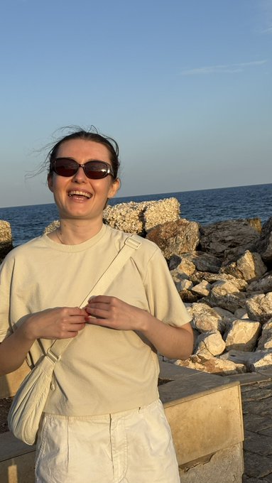 A smiling woman wearing sunglasses, a cream sweatshirt, and white pants poses by the sea at sunset with rocky breakwaters in the background.