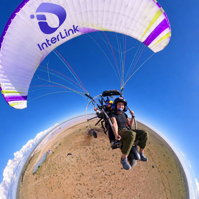 A 360-degree aerial view of a person paragliding with a white and purple 'InterLink' branded canopy over a dry landscape under a blue sky.