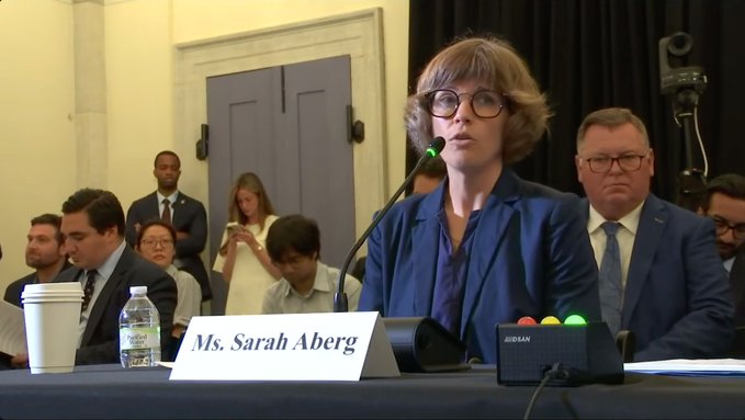 Ms. Sarah Aberg, identified by a nameplate, speaks into a microphone at a hearing or testimony, wearing glasses and a blue blazer.