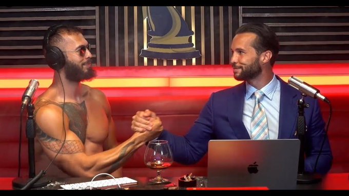 Two men shake hands across a table with microphones in a podcast studio, one shirtless with tattoos and headphones and the other in a blue suit, while a laptop displaying an Apple logo sits in front of them. The image promotes the $TATE token project by the Tate Brothers, whose crypto contract address is shown in the caption.