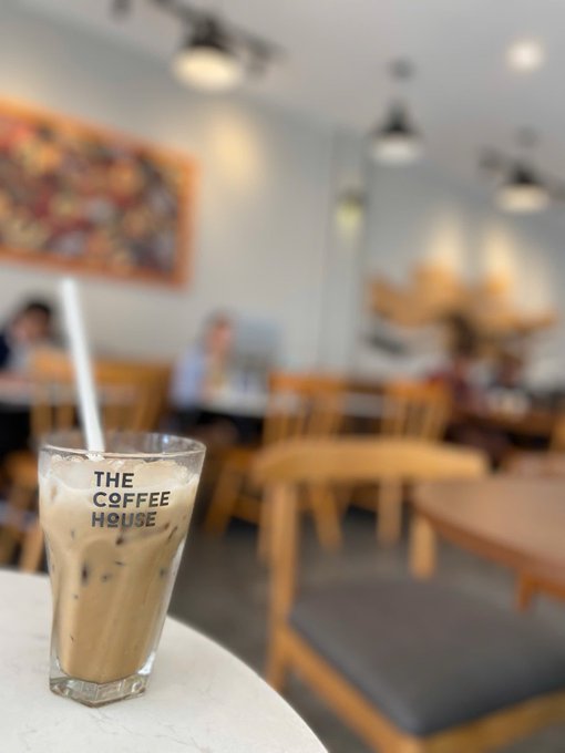 A glass of iced coffee with a straw sitting on a white table at 'The Coffee House,' with blurred cafe seating and patrons in the background.