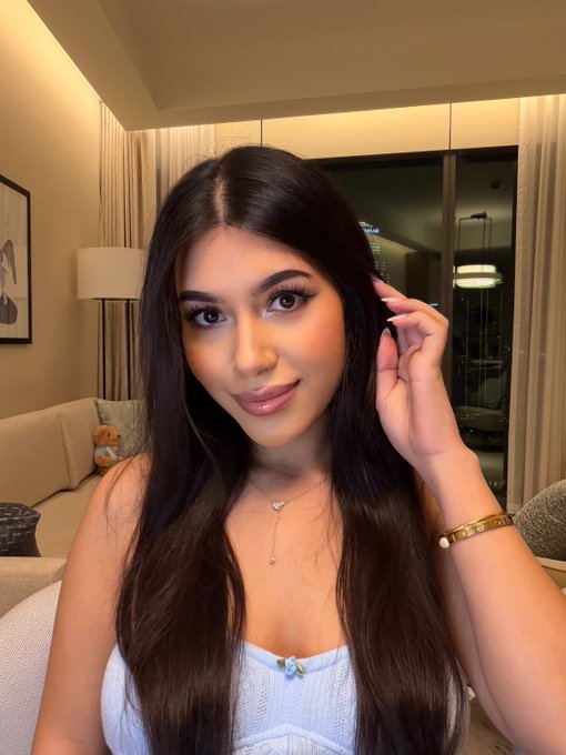 A selfie of a young woman with long dark hair wearing a light blue top and delicate necklace, posing indoors in a well-lit room with a lamp, couch, and city view through the window.