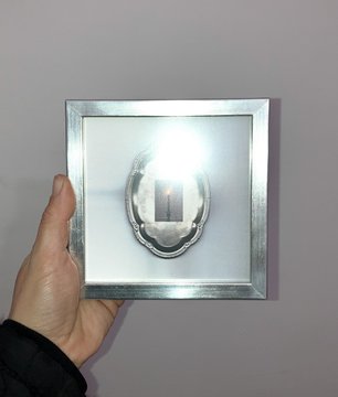 A hand holds a square metallic frame with a reflective center that appears to show a light or small object.