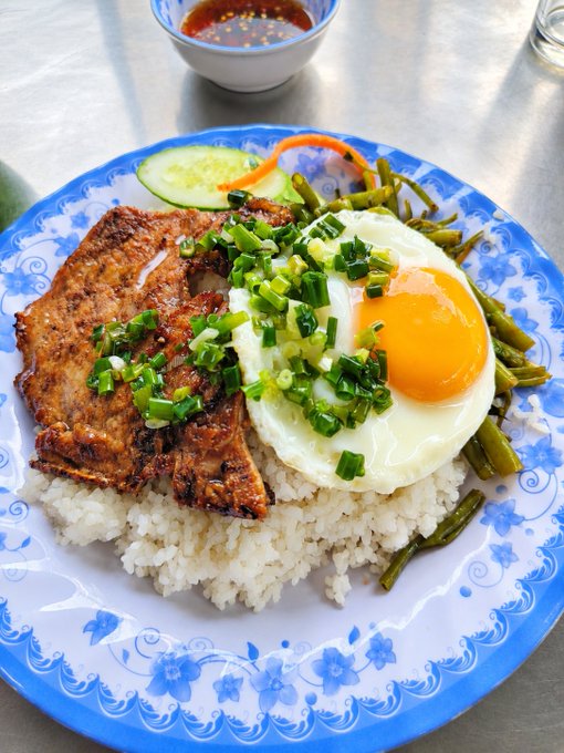 A plate of rice topped with grilled pork, a fried egg, chopped green onions, pickled green beans, and cucumber slices, with a small bowl of red chili sauce on the side.