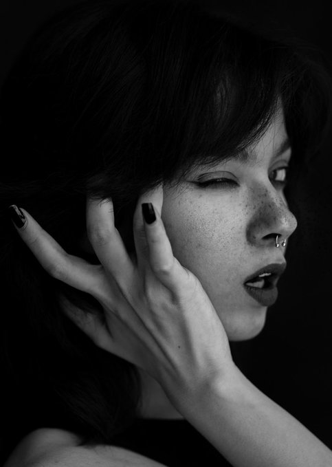 A high-contrast black and white portrait of a person with dark hair, a nose piercing, and dark nail polish, winking at the camera while resting their hand against their face.