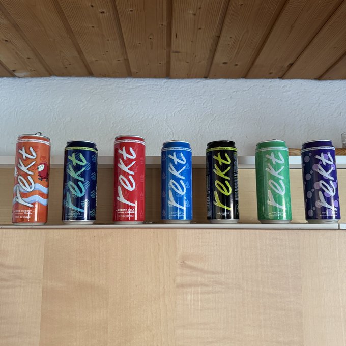 Seven colorful aluminum cans labeled 'rekt' energy drinks in orange, blue, red, blue, black, green, and purple flavors are lined up on a wooden shelf.