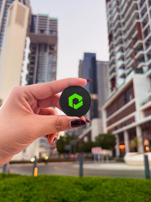 A hand with painted nails holding a black coin with a green hexagonal logo, set against a backdrop of modern high-rise apartment buildings in an urban setting.