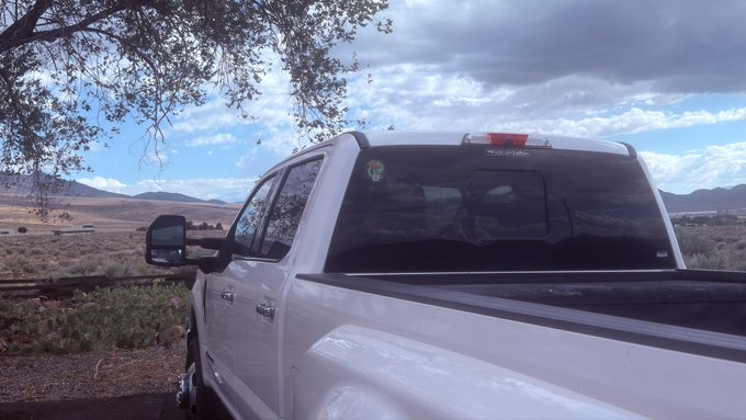 A rear view of a white pickup truck parked in a dry, open landscape with mountains in the distance under a partly cloudy sky.