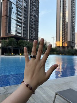 A hand is raised with the palm facing the camera, wearing a black ring and a red and black braided bracelet, with a blue swimming pool and tall apartment buildings in the background.