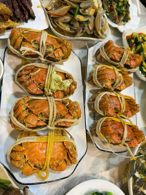 Multiple steamed crabs tied securely with ropes are served on rectangular plates, surrounded by other dishes like clams and vegetables.