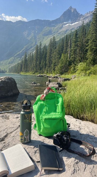 A bright green backpack, water bottle, camera, and open book are set on rocks by a serene lake with mountains in the background, making for a perfect "gm" to start a day of adventure.