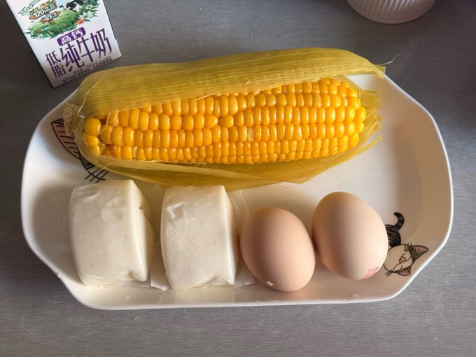 A white plate holds a yellow ear of corn, two white blocks of tofu in liquid, and two brown eggs, with a carton of milk partially visible in the upper left.