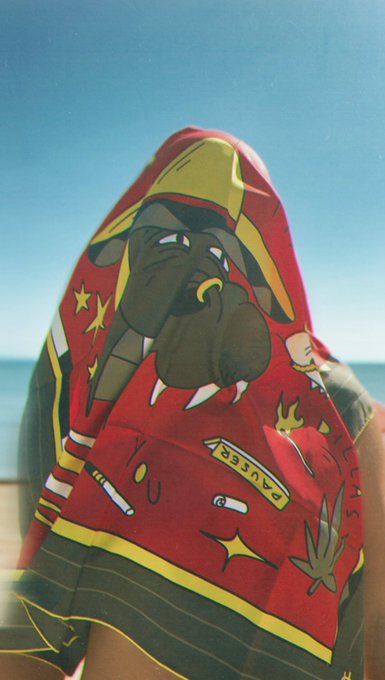 A photo of a person wrapped in a red graphic-printed towel/garment featuring a cartoon face, standing at a beach with blue sky and ocean visible behind them.