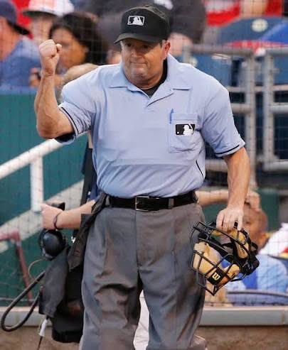A Major League Baseball umpire wearing a blue uniform and an MLB cap raises his fist while walking on the field holding his face mask.