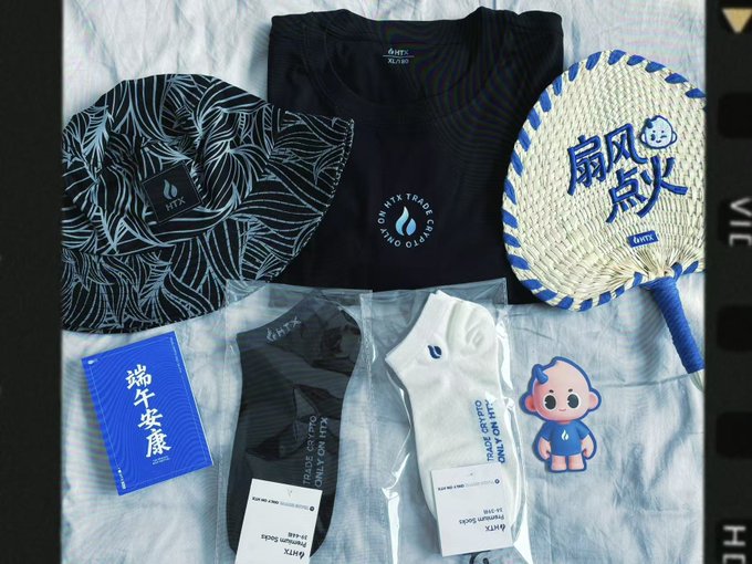 A flat-lay display of a Huobi HTX Dragon Boat Festival gift set featuring a black "HTX" t-shirt, a leaf-patterned hat, a woven palm-leaf fan with "扇风点艾" (Fan Wind, Light Moxa) embroidery, packaged socks, and a blue "端午安康" (Dragon Boat Festival Wishes) card, all arranged alongside a cartoon HTX mascot. The image illustrates the thoughtful, detail-oriented brand gift referenced in the caption, showcasing the palm-leaf fan and cultural touches the author praised.