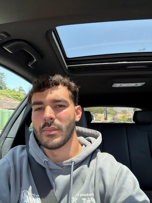 A selfie of a young man with curly brown hair and a beard sitting in the driver's seat of a car with the sunroof open on a sunny day.