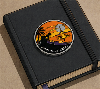 A circular pin or badge resting on a black notebook, depicting an orange sunset scene with a silhouette of a person playing basketball, accompanied by the name 'JOHN "Snap" KRAUS'.