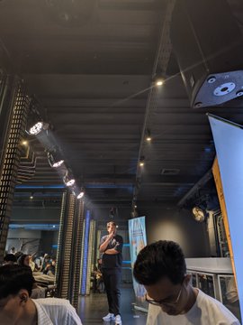 The image shows a man standing in front of a group of people who are sitting at tables with laptops, possibly attending an event or gathering. The man appears to be giving a speech or presentation while holding a microphone. There are several people seated around him, some using their laptops and others engaged in conversation. A few chairs can also be seen scattered throughout the scene.

The room has a dark atmosphere, which contrasts with the bright lights illuminating the area where the event is taking place. The presence of multiple people sitting at tables indicates that this gathering might be focused on technology or business-related topics.