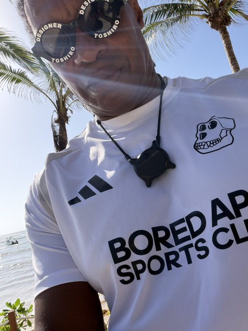 A man wearing sunglasses branded 'DESIGNER TOSHINO' and a white t-shirt with the text 'BORED AP SPORTS CL' and a skull logo, taking a selfie on a beach.