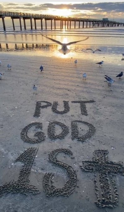 A beach scene at sunset with a pier in the background features the words 'PUT GOD 1st' written in the sand, as a seagull flies overhead and others stand nearby.