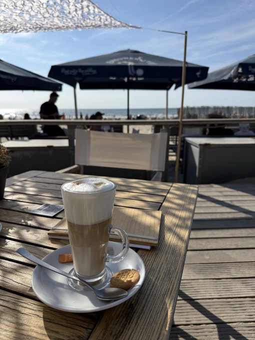 A layered latte with a cookie served on a wooden table at a beachside cafe, with umbrellas, the sea, and sky visible in the background.