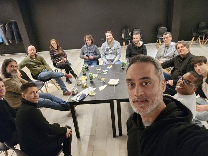 A group selfie of about a dozen people sitting around a conference table with refreshments in a modern meeting room.