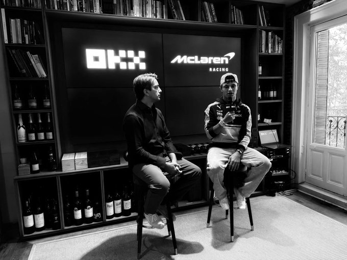 Two men sit on stools in a library-style room during what appears to be an interview, with illuminated "OKX" and "McLaren Racing" logos displayed on the wall behind them, flanked by bookshelves and wine bottles. The setting captures the convergence of cryptocurrency (OKX) and motorsports (McLaren Racing) mentioned in the post, with one man in a racing team polo suggesting a McLaren driver is being interviewed alongside an OKX representative.