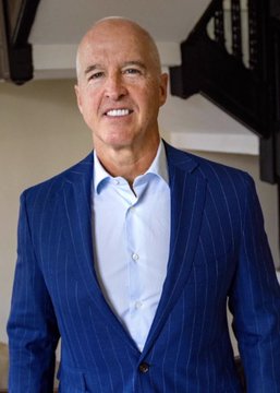 A smiling man in a blue pinstripe suit and light blue dress shirt stands indoors near a staircase, identified as Mike Minogue, the projected winner of the Massachusetts Republican gubernatorial primary who is set to challenge Governor Maura Healey in the November election.