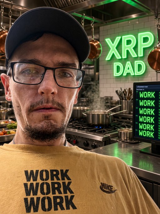 A man wearing glasses, a black cap, and a yellow "WORK WORK WORK" Nike t-shirt stands in a kitchen, with a neon "XRP DAD" sign and a monitor displaying "WORK" on a green background.