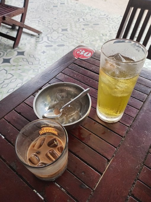 A photo of two drinks on a wooden slatted table - a glass of iced brown coffee and a glass of iced yellow tea, accompanied by a small metal bowl with a spoon, on a table marked '30'.