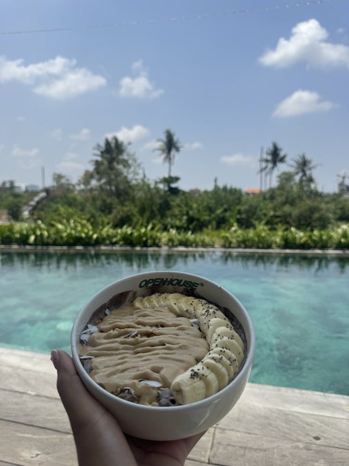 A hand presents a smoothie bowl from "OPENHOUSE" topped with sliced bananas and drizzles of peanut butter, set against a stunning backdrop of a turquoise infinity pool and tropical palm trees in Bali.