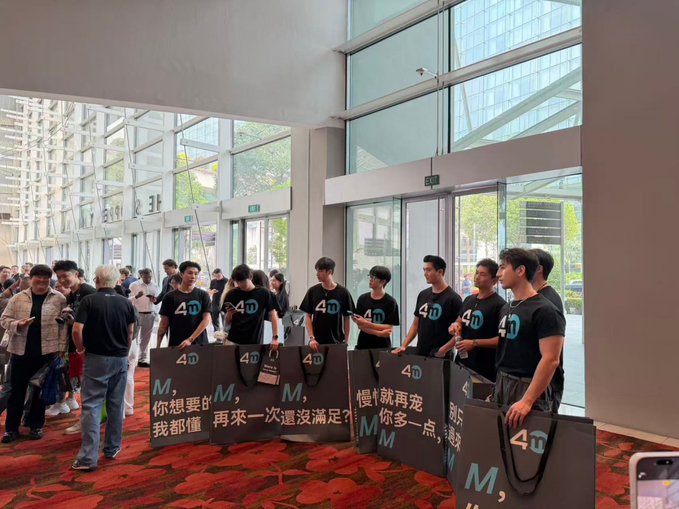 A group of men wearing matching black t-shirts with '4D' branding stand in a lobby holding promotional signs with Chinese text and 'M' logos, appearing to be at an event.
