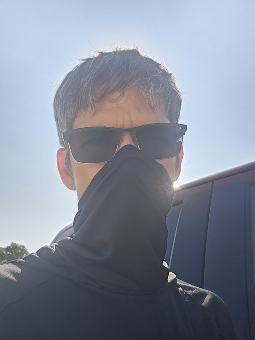 A young man wearing sunglasses and a black neck gaiter pulled up over his face takes a selfie outdoors under a clear blue sky, with sun flare peeking out from behind a dark vehicle. The image pairs with the "Gm" crypto greeting, visually representing the mysterious, low-profile vibe often associated with early-morning crypto traders and Cardano ($ADA) holders.