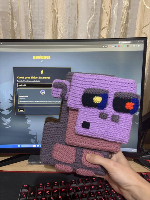 A hand holding a crocheted purple and pink robot plushie in front of a computer monitor displaying the BitBoy x site for checking Bibit list status.