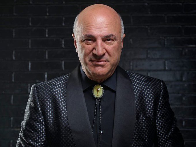 A portrait of a bald older man wearing a patterned black jacket and bolo tie with a green stone, photographed against a black brick wall.