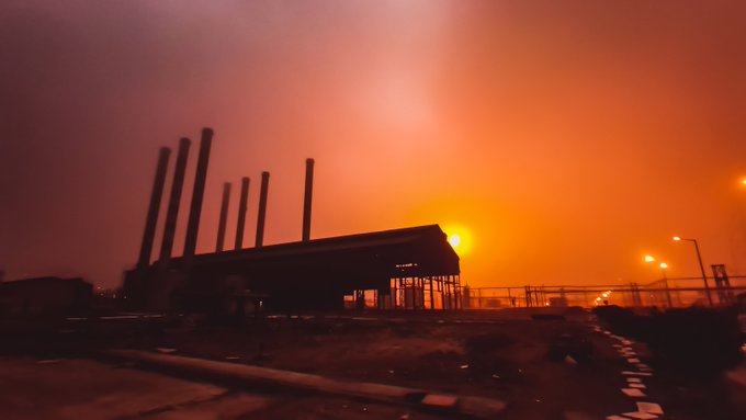 A silhouette of an industrial facility with smokestacks stands against a dramatic, bright orange and pink hazy sky at sunset.