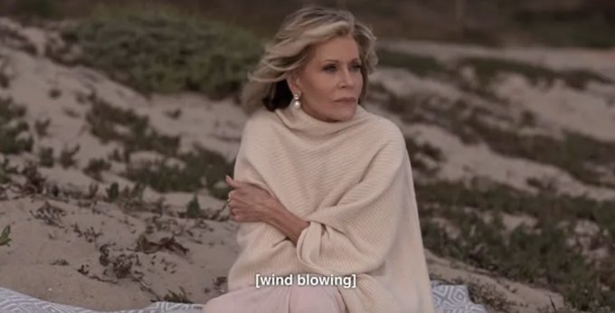 A blonde woman wrapped in a cream-colored blanket sits outdoors on a beach setting, with a caption reading '[wind blowing]' indicating wind effects in the scene.