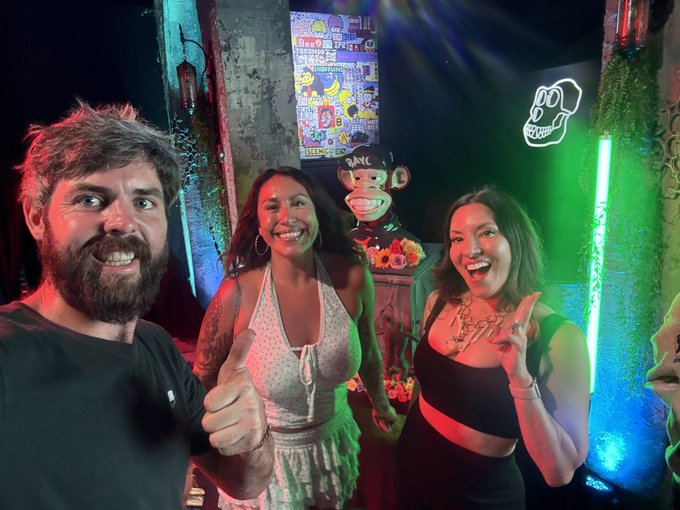Three people smiling and posing at what appears to be a nightclub or bar, with one giving a thumbs-up and another making a peace sign, with a mischievous monkey statue visible behind them.