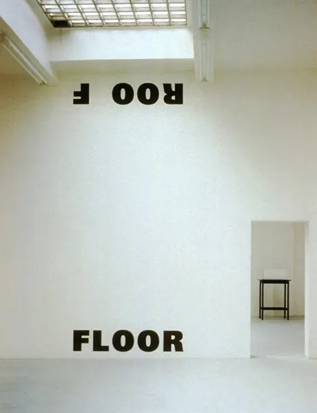 An art installation in a white gallery space displays the word 'FLOOR' correctly oriented on the lower wall, while the same word 'FLOOR' appears upside down on the upper wall near the ceiling, questioning the perception of ceiling and floor.