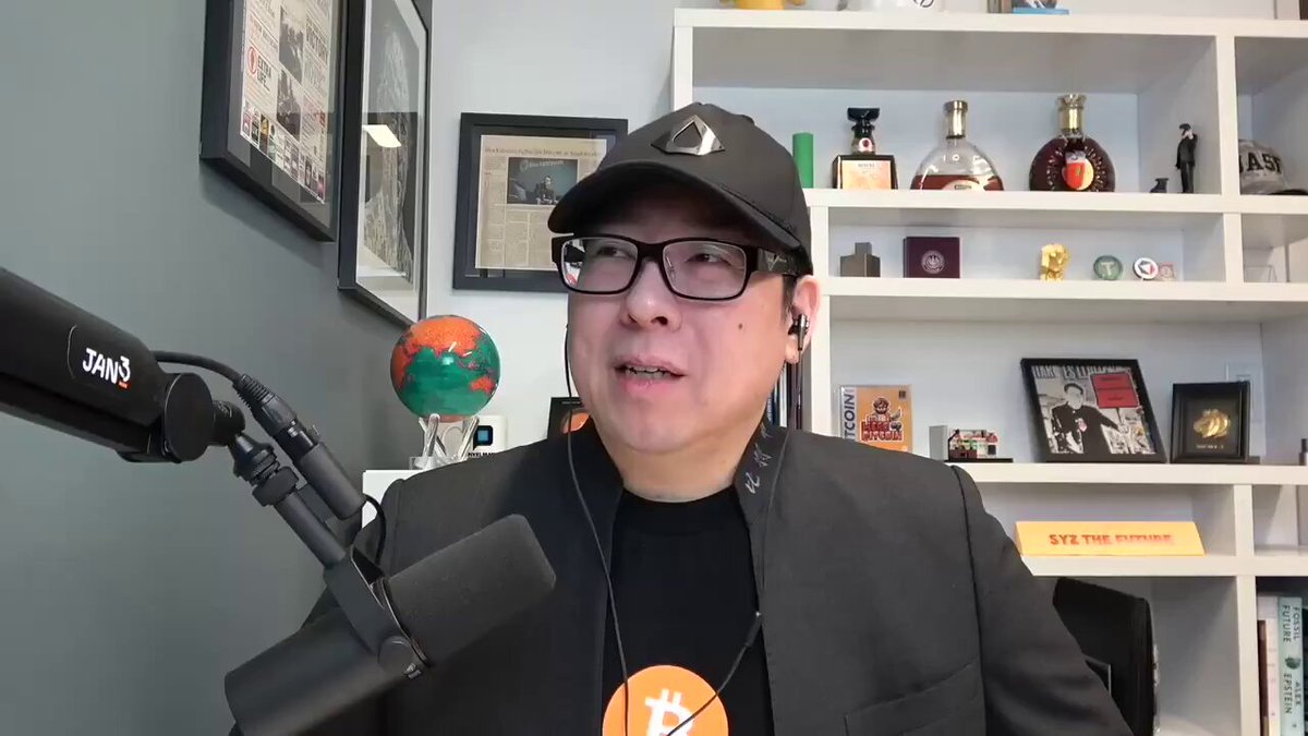 The image features a man wearing glasses, a black hat, and a suit talking into a microphone. He appears to be discussing Bitcoin or another cryptocurrency-related topic. The background of the image shows a bookshelf filled with various items such as bottles, books, and other objects. There is also an orange globe in the scene.