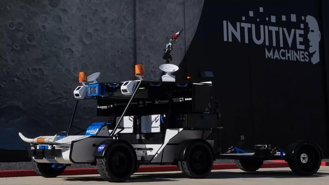 The image features a large, black truck with two levels of antennas mounted on top. The truck has a trailer attached to its back that also has an antenna. In front of this truck, there's a smaller vehicle, possibly a car or a bus, which is parked next to it. The background of the image shows a building and a wall, but the main focus remains on the two-leveled truck with antennas.