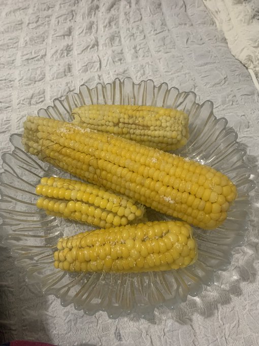 A clear glass fluted dish holding four pieces of cooked yellow corn on the cob, sprinkled with seasoning, set on a white textured tablecloth.