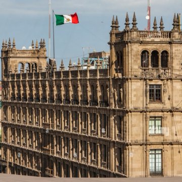 A historic stone building with Gothic architectural details and spires, with a Mexican flag flying on top against a cloudy sky.