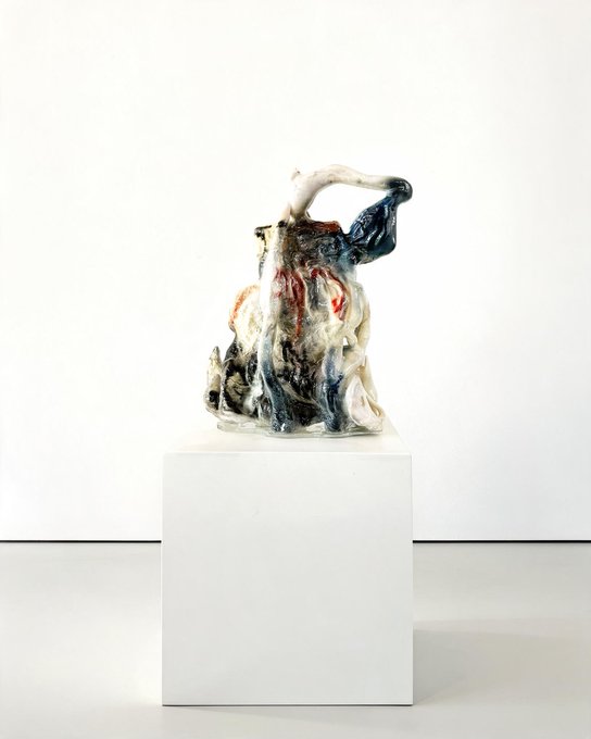 A glazed ceramic sculpture with abstract, distorted forms in white, blue, and red sits atop a white pedestal in a gallery space.
