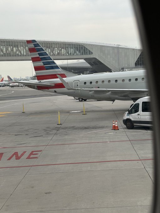 A view from inside an aircraft window showing the tail and rear fuselage of an American Airlines plane with its signature red, white, and blue striped livery, parked on the tarmac near a terminal with jet bridges.