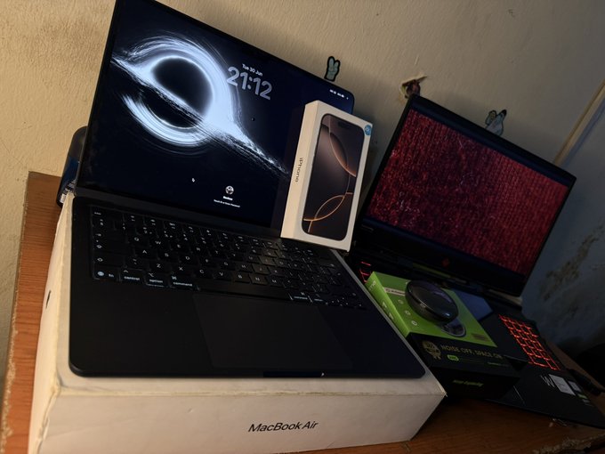 A MacBook Air box serves as a stand for an open laptop displaying a black hole wallpaper with the time "21:12," alongside an iPhone box, a red-backlit keyboard, mouse, and external monitor—visually capturing six months of dedicated tech grinding and setup investment.