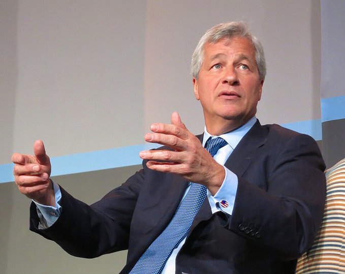 A gray-haired man in a dark suit and blue tie gestures with both hands while speaking, appearing to be at a panel or conference.
