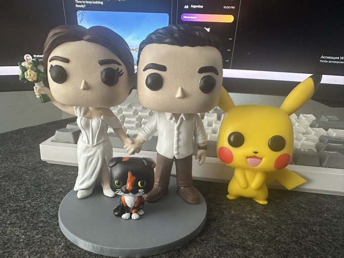 A custom Funko Pop-style wedding figurine shows a bride and groom in white and beige attire, accompanied by a Pikachu figure and a small calico cat, displayed on a desk in front of a keyboard and monitor.