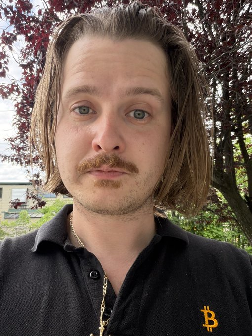 A young man with long brown hair, a mustache and goatee, and striking blue eyes poses outdoors in front of a tree with dark red foliage, wearing a black polo shirt with a Bitcoin logo. The image personalizes the post, putting a face to a stressed, debt-burdened pharmacist who is holding out hope that a Bitcoin price drop to 10k would bring financial relief before committing further to NFTs.